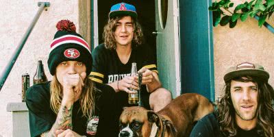 Three members of band Dune Rats sitting outside a house on the steps, drinking beer, with a boxer dog, wearing caps and a San Francisco beanie