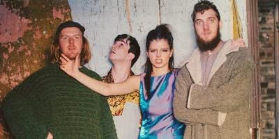 Female member of The Jungle Giants holds bandmates chin, while they pose in front of a rustic wall