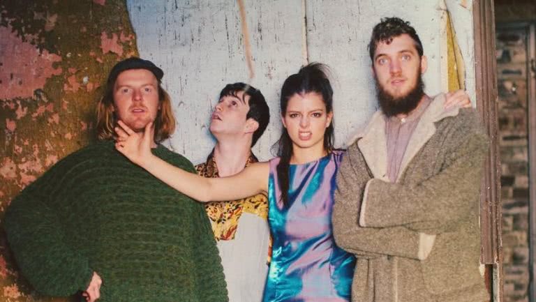 Female member of The Jungle Giants holds bandmates chin, while they pose in front of a rustic wall