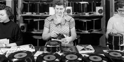 Black and white vintage photo of three women working in a vinyl pressing factory, packaging 7" singles into record sleeves
