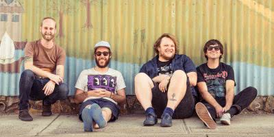 Four members of Smith Street Band, Wil Wagner second from right, sit on the footpath in front of a mural painted on a corrugated metal fence