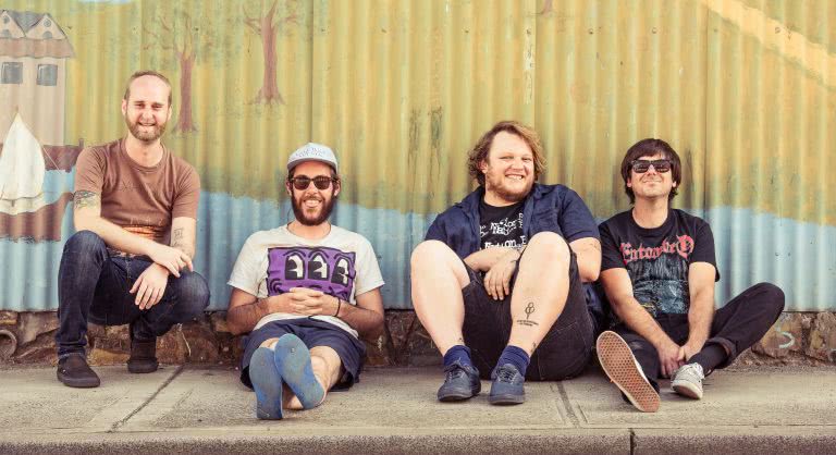 Four members of Smith Street Band, Wil Wagner second from right, sit on the footpath in front of a mural painted on a corrugated metal fence