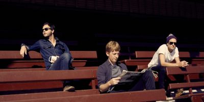 The three members of Fountaineer, one reading a newspaper, sitting in the stands of a suburban sportsfield