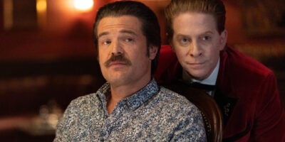 A man in a red velvet jacket leans beside a seated man in a scene from The Highest Stakes.