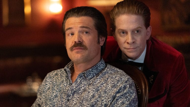 A man in a red velvet jacket leans beside a seated man in a scene from The Highest Stakes.
