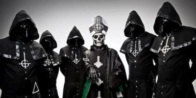 5 men standing in hooded costumes with faces covered and man standing in the middle with a skull mask in a black cloak