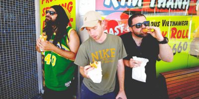 Three men standing in front of a shop window eating take away food