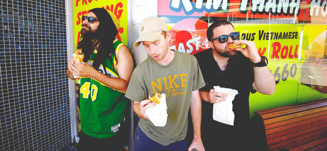 Three men standing in front of a shop window eating take away food