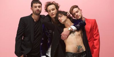 Four men standing in front of a pink background. One in a red jacket and three in black jackets, man standing infant has no shirt on