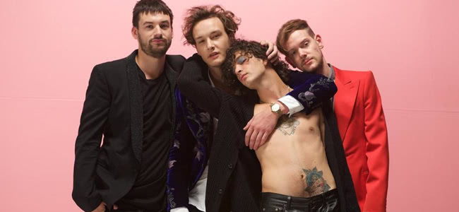 Four men standing in front of a pink background. One in a red jacket and three in black jackets, man standing infant has no shirt on