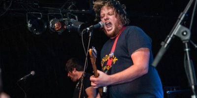 Man singing into microphone on stage playing guitar with navy t-shirt on