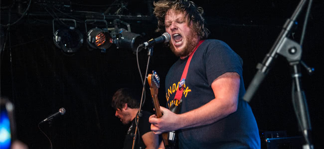 Man singing into microphone on stage playing guitar with navy t-shirt on