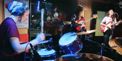 Three band members playing drums and guitar wearing a beanie and red and black striped t-shirt