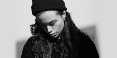 Tash Sultana looking down in black top and black beanie.