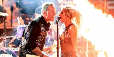 Man and woman sining into microphone on stage with fire burning behind them