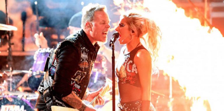 Man and woman sining into microphone on stage with fire burning behind them