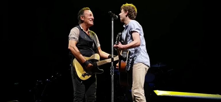 Bruce Springsteen playing guitar sining on stage with boy in light blue shirt playing guitar