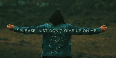 Man standing in a green field wearing a denim hoodie with his back to the camera and arms stretched out with writing across his arm span