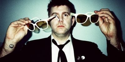 Man in black suit, whist shit and black tie holding two pairs of white sunglasses with a tattoo on his write looking at the camera