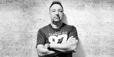 Black and white image of Peter Hook standing against a wall with his arms crossed