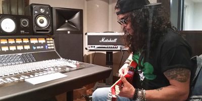 Slash wearing a backwards cap, playing a guitar sitting in a recording studio