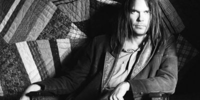 Neil Young sitting on a chair with a dark jacket on looking at the camera