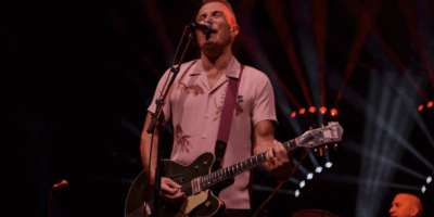 James Reyne on stage singing into a mic playing guitar