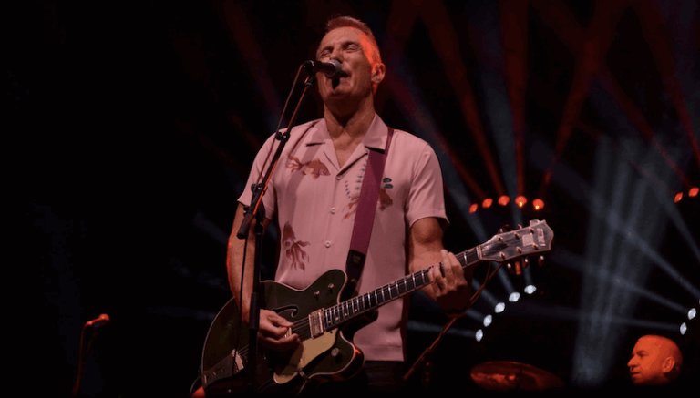 James Reyne on stage singing into a mic playing guitar