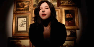 Lucy Dacus standing in front of a gallery wall with a spotlight on her, staring down the camera