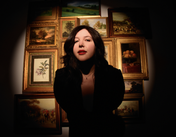 Lucy Dacus standing in front of a gallery wall with a spotlight on her, staring down the camera