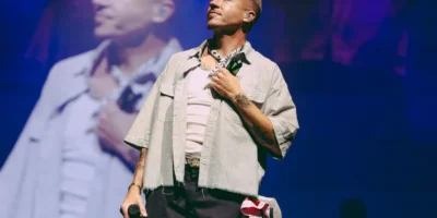 Macklemore holds a microphone at his side with one hand over his chest, a large screen showing him behind.