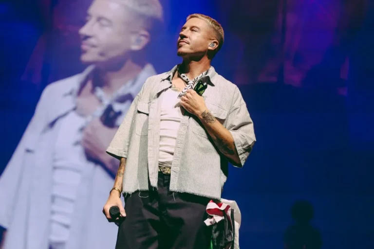 Macklemore holds a microphone at his side with one hand over his chest, a large screen showing him behind.